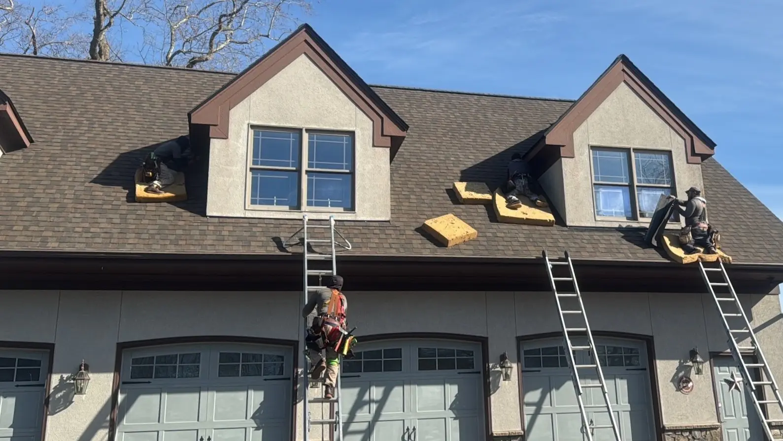 Workers install yellow foam insulation on a dormer roof of a two-story house, using ladders and safety gear.