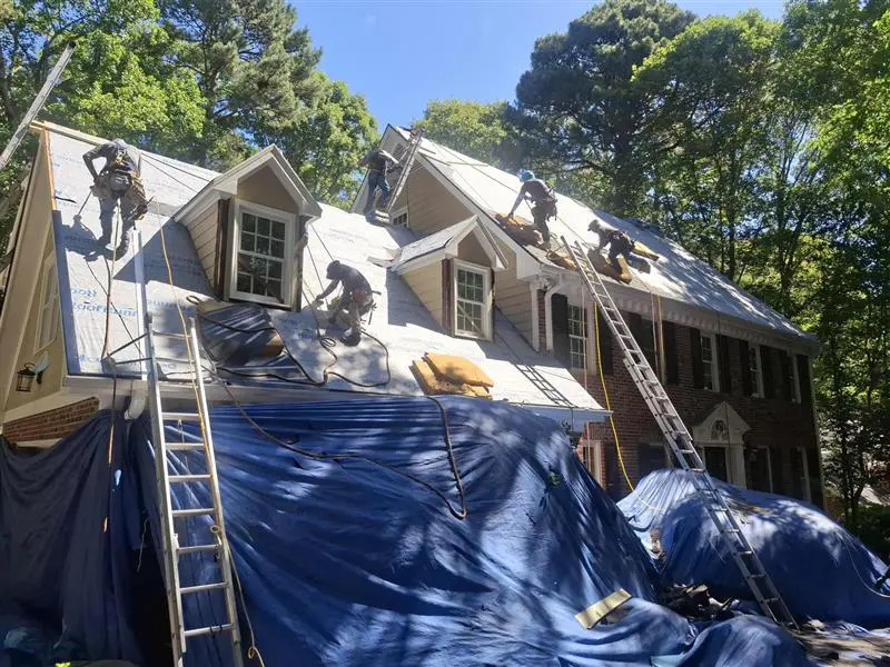 House with workers repairing a slate roof; blue tarps cover the yard and several ladders lean against the roof.