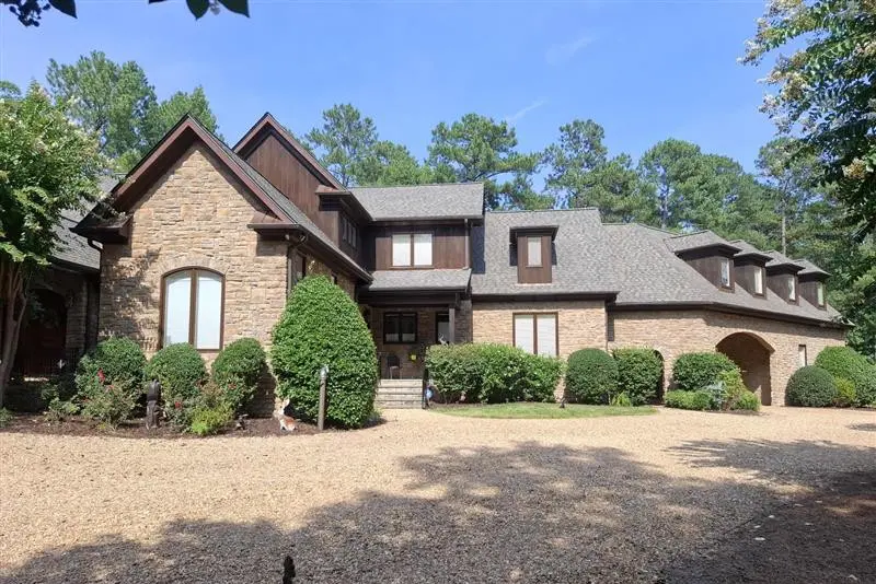 Front view of a large rustic two-story house with stone and wood siding, peaked roofs, dormer windows, and neatly trimmed shrubs along a gravel driveway.