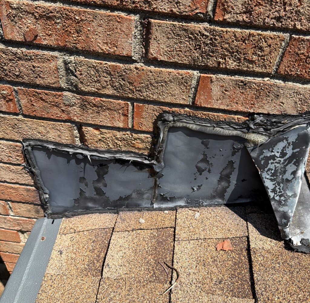 Close-up of a brick wall with damaged roof flashing and ripped, peeling roofing material at the lower edge.